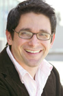 A smiling man with dark hair and glasses wearing a light pink shirt and dark jacket.