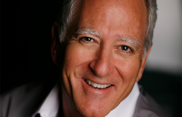 Close-up portrait of a smiling man with gray hair and eyebrows wearing a white shirt and dark jacket.