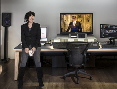 A woman in a black blazer stands in a recording studio with a mixing console and monitors.