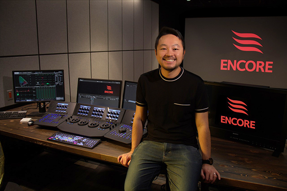 A man in a black t-shirt sits in a studio with multiple monitors and video editing consoles.