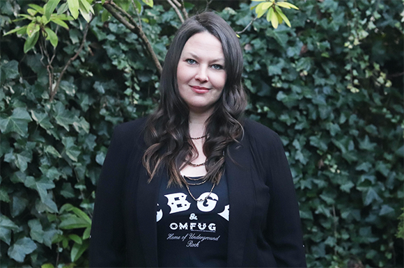 A woman with long dark hair wearing a black blazer and a black graphic t-shirt stands before green ivy.