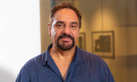 A man with dark hair and a goatee wearing a blue button-down shirt looking at the camera.