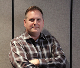 A man with short brown hair wearing a brown and white plaid shirt, standing with arms crossed.