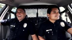 Two male police officers in dark uniforms sitting in the front seats of a patrol car, smiling.