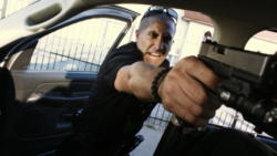 A man in a dark uniform aims a handgun toward the camera from inside a car.