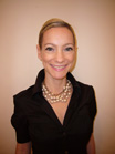 A smiling woman with blonde hair wearing a black collared shirt and a multi-strand pearl necklace.