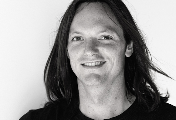 A smiling man with long, dark hair looking directly at the camera against a plain background.
