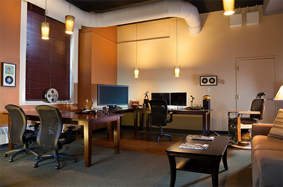 A media studio with desks, computer monitors, film reels, and office chairs under warm hanging lights.
