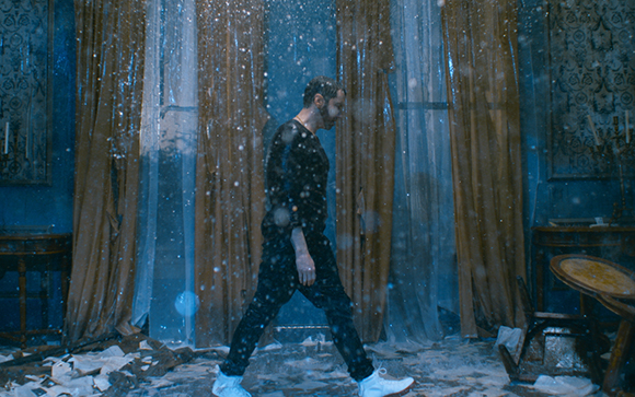 A man in dark clothing walks through falling snow inside a room with long, heavy curtains.
