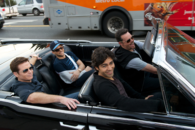 Four men smiling and wearing sunglasses while riding in a black convertible on a city street.