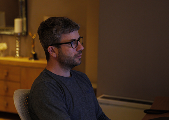 A man with salt-and-pepper hair and glasses looking off-camera in a dimly lit room.