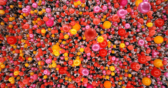 A large crowd of people releasing a dense mass of red, orange, pink, and yellow balloons.