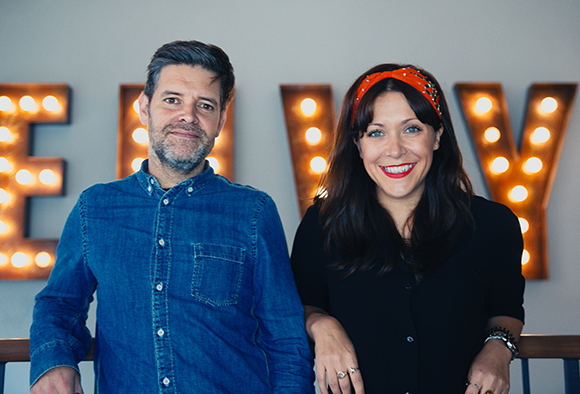 A man in a blue denim shirt and a woman in a black top smile in front of glowing marquee letters.