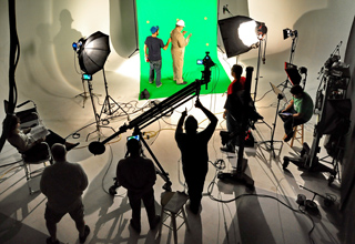 A film crew operates cameras and lighting around two actors in front of a bright green screen.