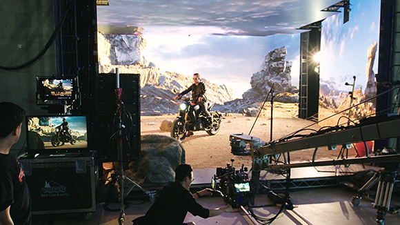 A film crew uses a camera crane to film a person riding a motorcycle against a large digital landscape screen.