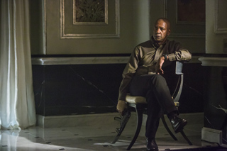 A man in a dark shirt and trousers sits in a chair, holding a handgun in a dimly lit room.