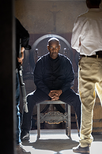 A man in a dark jacket sits on a chair while two people stand in the foreground, partially obscuring the view.