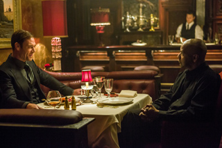 Two men in formal attire sit across from each other at a white-clothed table in a dimly lit restaurant.
