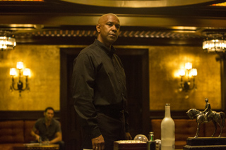 A man in a black button-down shirt stands in a dimly lit, gold-toned room with sconces.