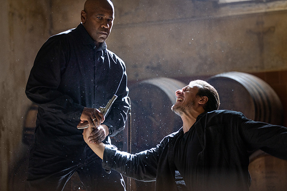 A man in a black jacket holds a handgun to the head of a man recoiling in a dusty, dark room.