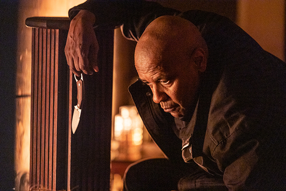 A man in dark clothing leans against a wooden pillar, holding a small knife by its handle.