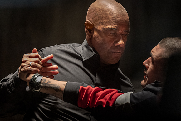 A bald man in a black shirt grips the arm of a younger man during a tense confrontation.