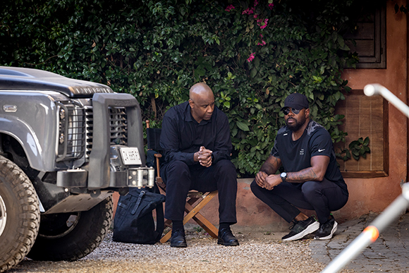 Two men in dark clothing sit on chairs outdoors next to a large grey off-road vehicle.