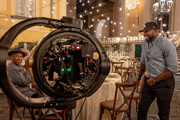 A camera operator films a man in a hat sitting at a decorated dinner table outdoors at night.
