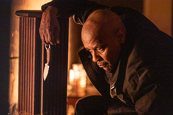 A bald man in dark clothing crouches, holding a knife pointed downward near a wooden pillar.