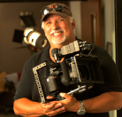A smiling man with gray hair and a goatee holding a large professional film camera.