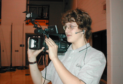 A person with glasses and a headset holding a large video camera.