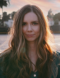 A woman with long, wavy brown hair looks toward the camera against a soft sunset background.