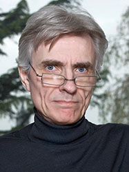 A man with grey hair and glasses wearing a black turtleneck against a blurred outdoor background.
