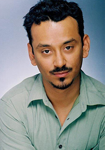 A man with dark, wavy hair and a goatee wearing a light green collared shirt against a blue background.