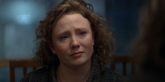 A woman with wavy reddish-brown hair looking down with a sorrowful expression.