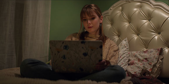 A woman with reddish hair sits cross-legged on a bed, looking down at a floral patterned laptop.