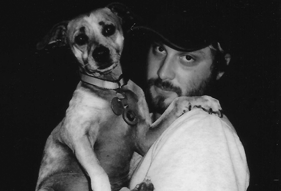 Black and white photo of a man with a beard and cap holding a small dog with a collar.
