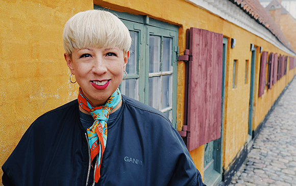 A woman with short blonde hair and a blue patterned scarf smiles in front of a yellow building.