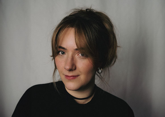 A woman with light brown hair, freckles, and a nose ring looks toward the camera against a neutral background.