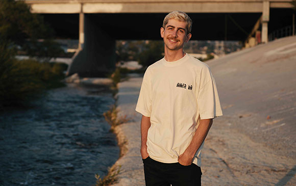 A smiling young man with blonde hair wearing a white t-shirt stands by a concrete canal under a bridge.