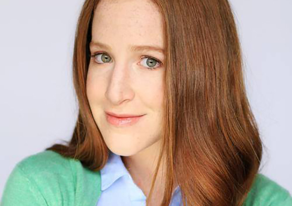 A woman with long reddish-brown hair and green eyes wearing a light blue shirt and green cardigan.