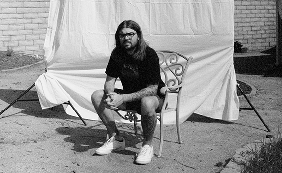 A man with long hair and glasses sits in a metal chair before a large white sheet outdoors.