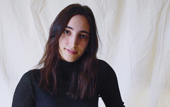 A woman with long dark hair and a black turtleneck poses against a white wrinkled fabric background.