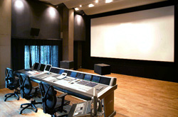 A dark media editing room featuring a long control desk with multiple monitors and a large white projection screen.
