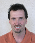A man with dark curly hair and a goatee smiling while wearing an orange collared shirt.