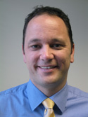 A smiling man with short dark hair wearing a blue button-down shirt and a gold necktie.