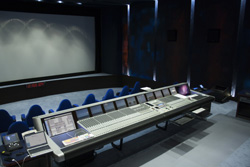 A long, professional audio mixing console sits in a dark studio before a large projection screen.