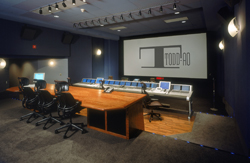A dark editing suite featuring a long wooden desk with multiple monitors and a large projection screen.