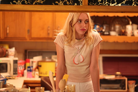 A blonde woman in a light pink graphic t-shirt looks off-camera in a warm-toned kitchen.