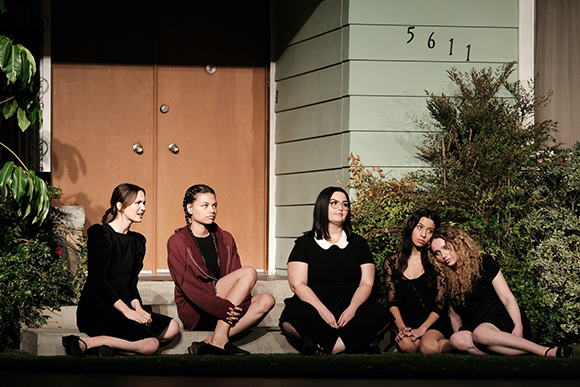 Five women in dark clothing sit on outdoor steps in front of a light green house.